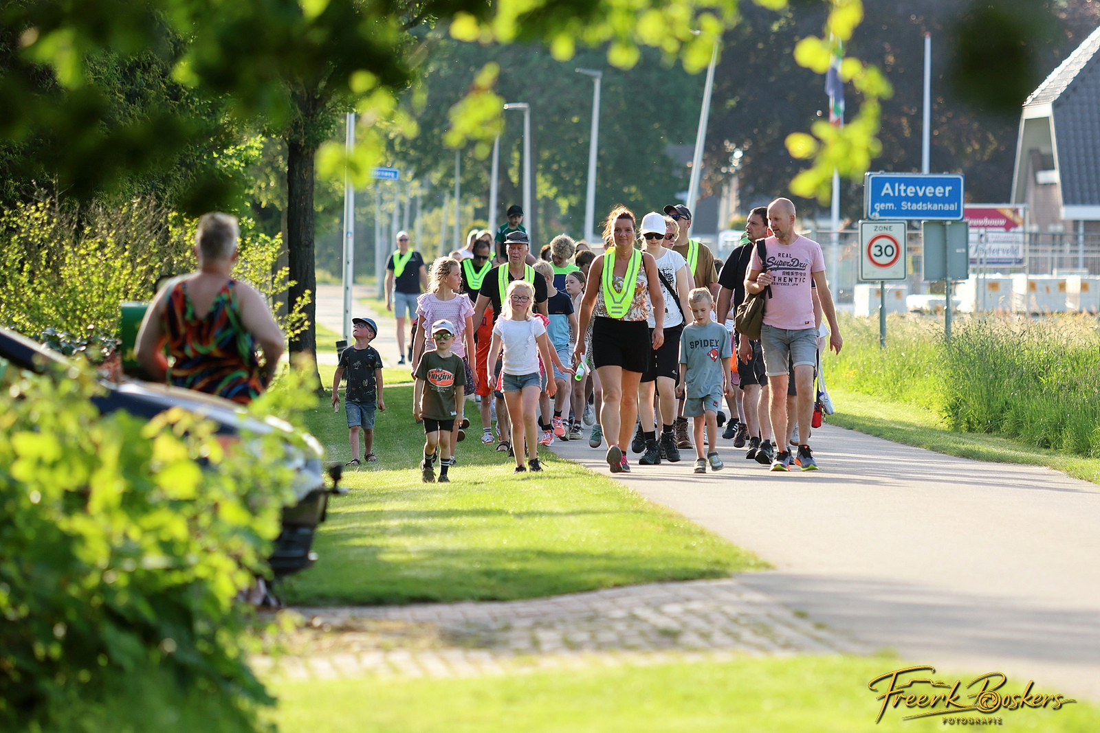 Groep wandelaars met kinderen loopt langs een weg, met groene hesjes en verkeersborden op achtergrond.